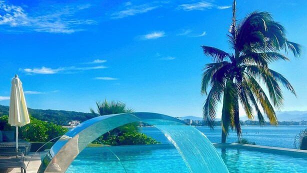 A clear blue pool with a waterfall feature, surrounded by palm trees and a clear sky.