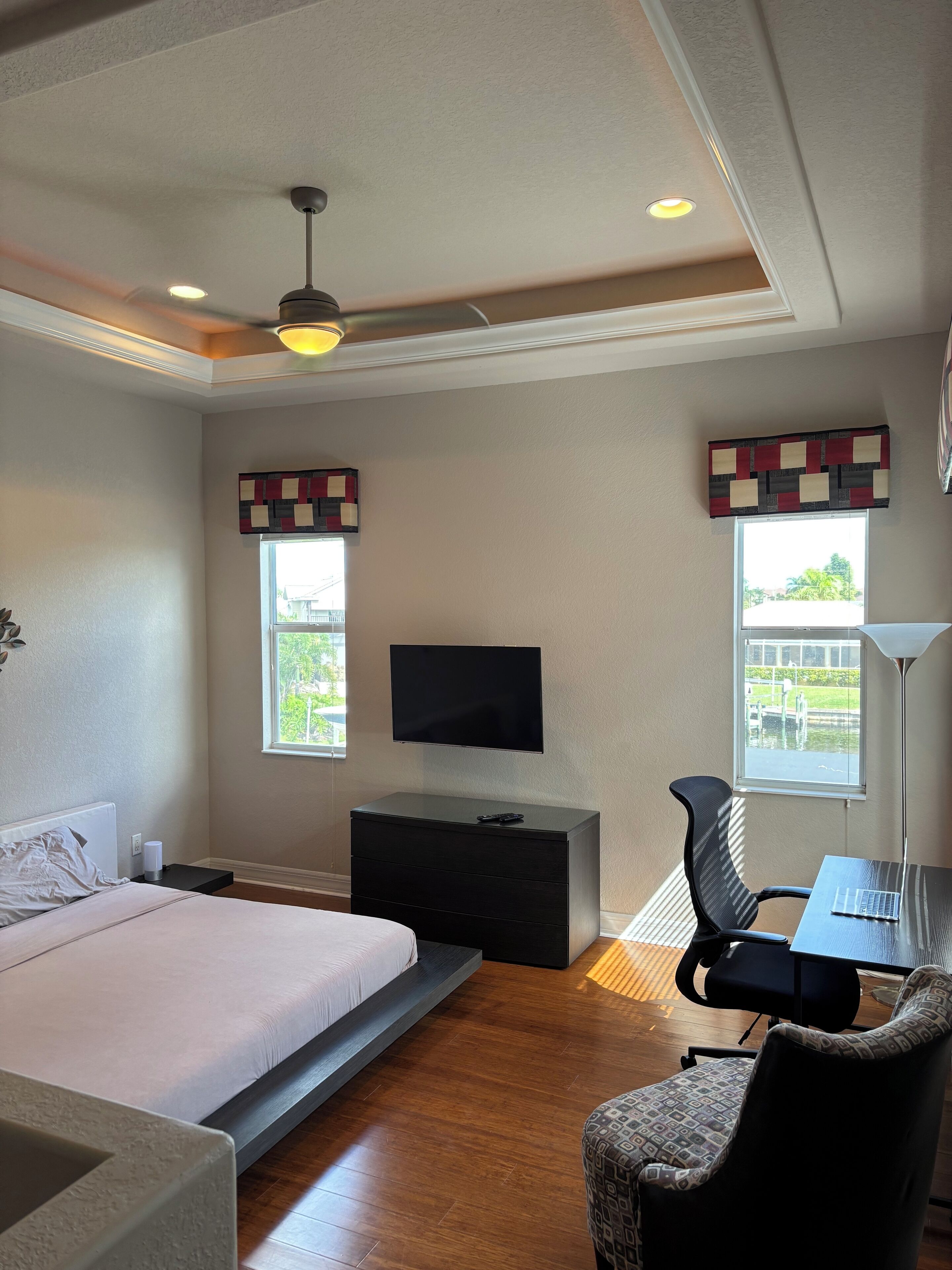 A bedroom with a bed, a desk, a chair, a TV, and two windows with curtains.