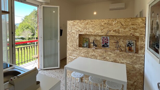 Una cucina moderna con un piano di lavoro bianco, sgabelli da bar, un muro di legno decorato con opere d'arte e una porta scorrevole in vetro che dà su un balcone.