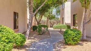 A pathway flanked by neatly trimmed bushes and trees, leading to a building with a beige facade and windows. - Modern Condo Near Stadiums & Westgate Fun (Phoenix)