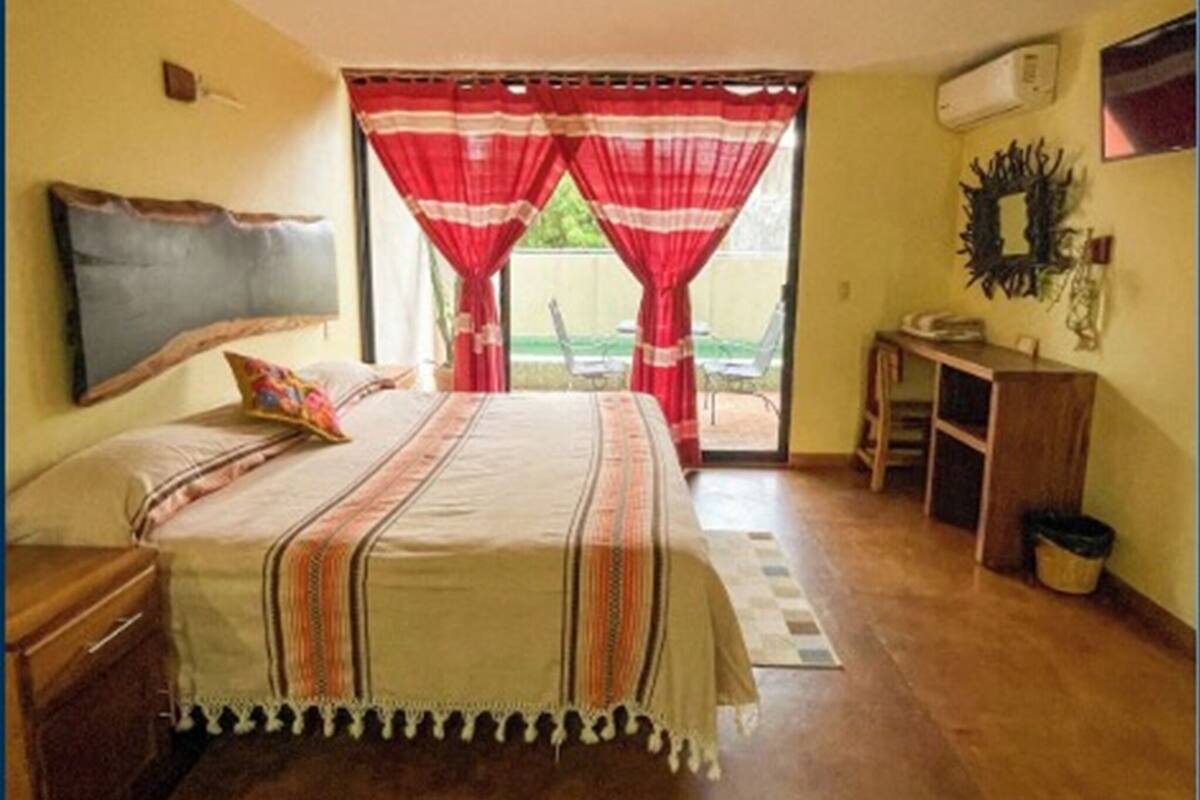 Une chambre à coucher avec un lit, une table de chevet, un bureau et une chaise. Il y a une fenêtre avec des rideaux rouges et une vue sur l’extérieur.
