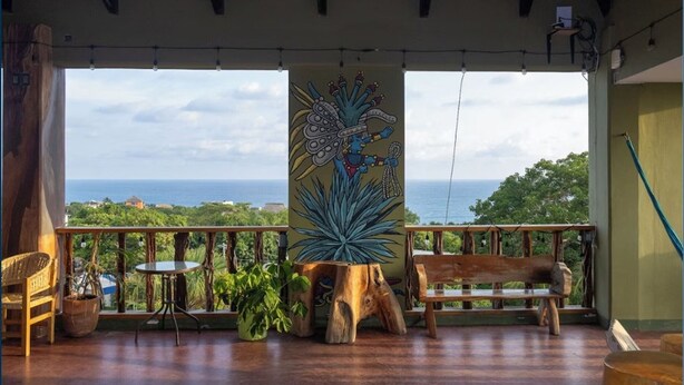 Un balcon avec vue sur la mer, un banc en bois, une table et une grande fresque murale.