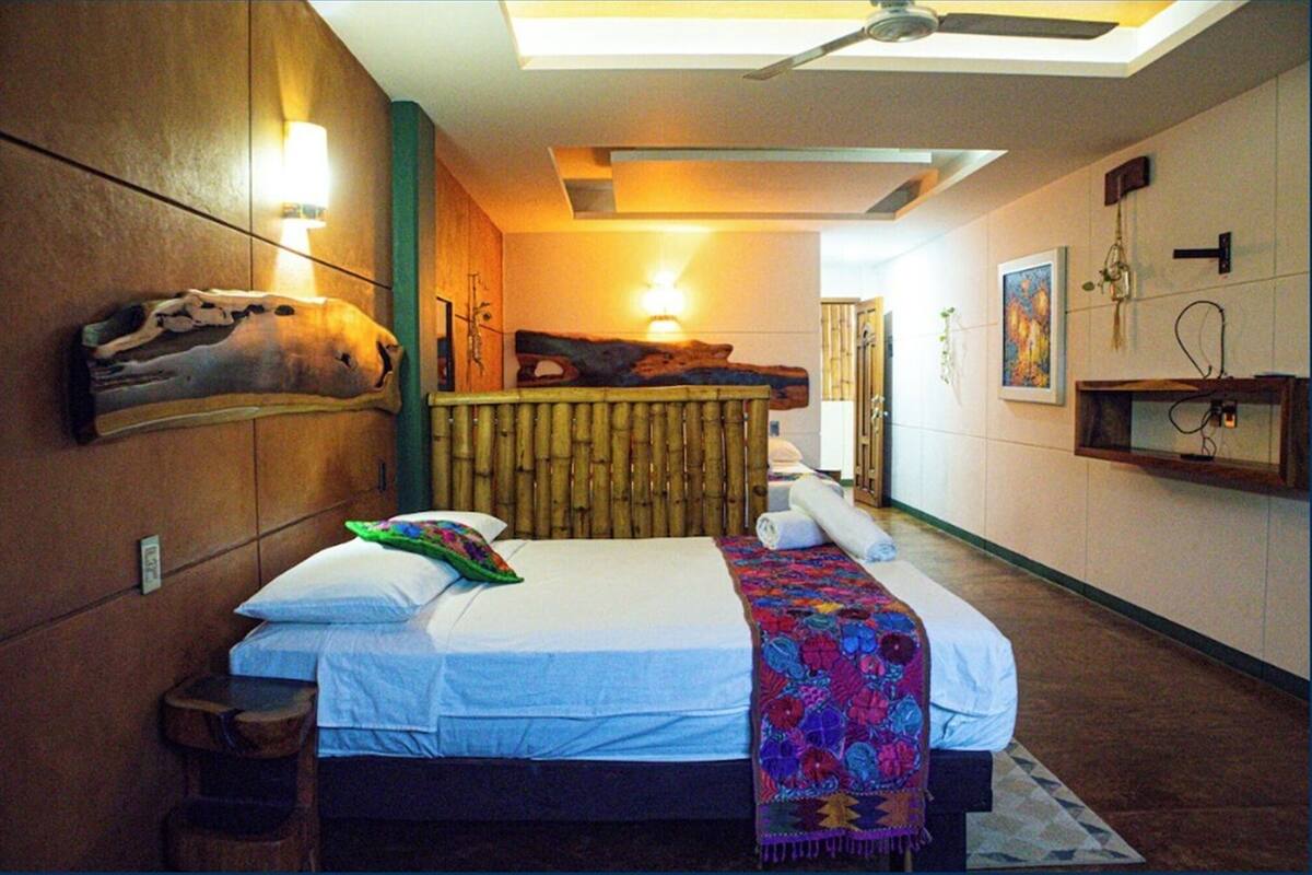 Une chambre d’hôtel moderne avec un grand lit, un ventilateur de plafond, une tête de lit en bois et une étagère murale avec une lampe.