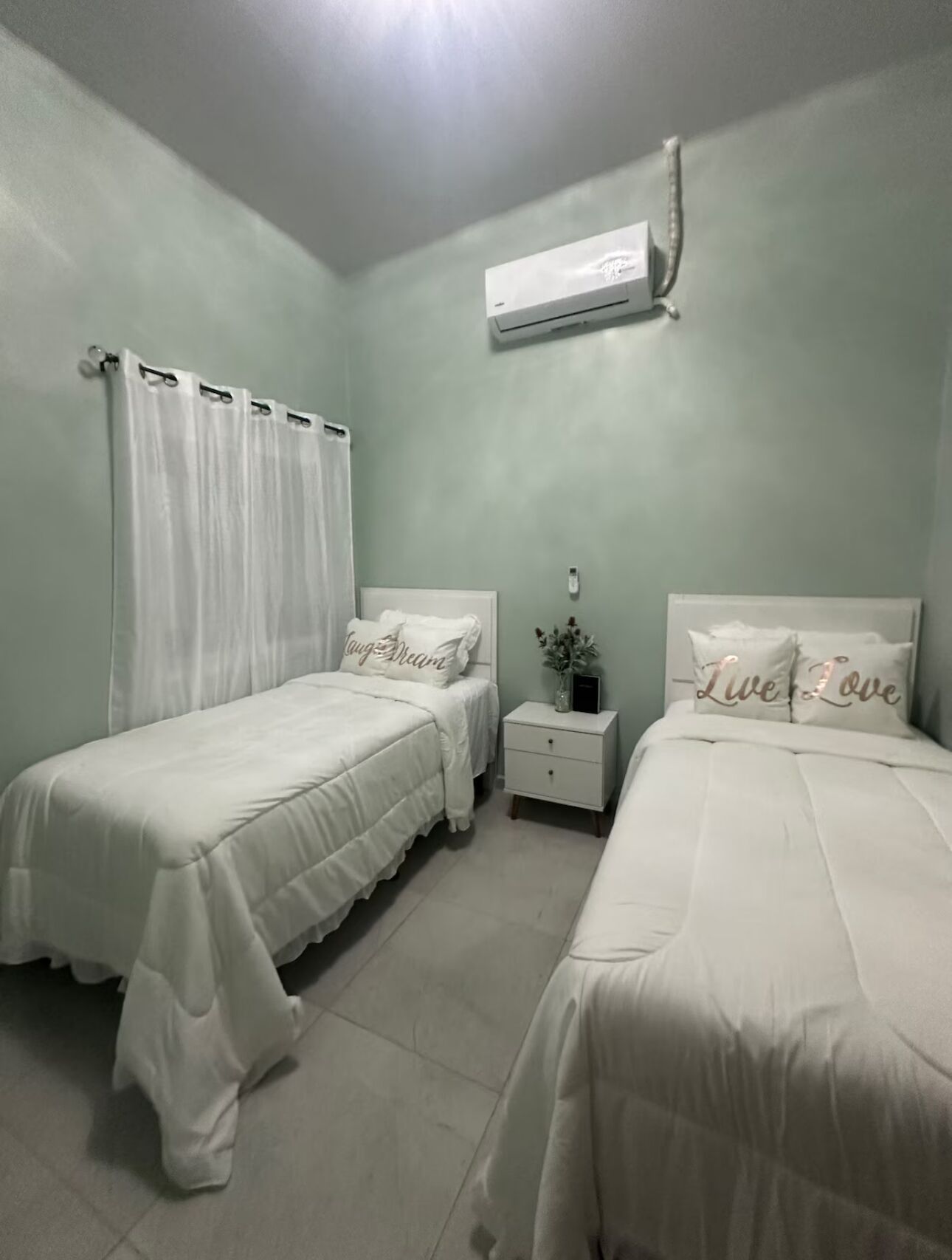 Quarto com duas camas, roupa de cama branca, mesa de cabeceira com um vaso e um aparelho de ar condicionado na parede.