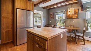 A kitchen with wooden cabinets, a stainless steel refrigerator, a wooden island with drawers, a dining table with chairs, and a window with curtains.