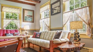 A cozy living room with a sofa, a wooden coffee table, and a rug. There are two windows with curtains, a lamp, and framed pictures on the wall.