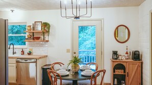A dining area with a round table, chairs, a chandelier, a kitchen with a dishwasher, and a view through a door. - 5 Minutes To Dollywood - Cottage With Fire Pit, Coffee Bar & Private Deck (Pigeon Forge)