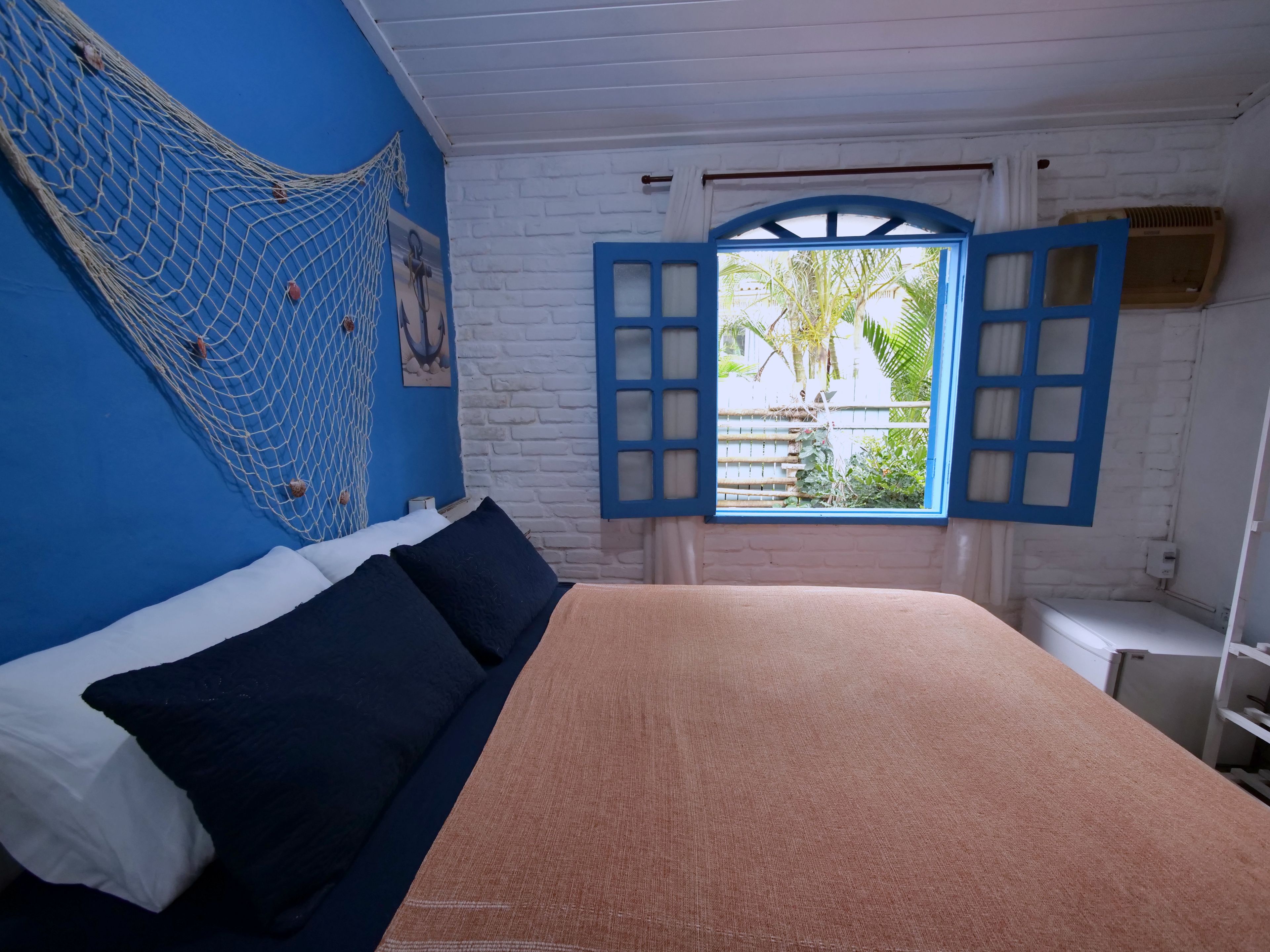 Een kamer met een bed, een hangmat, een raam met blauwe luiken en uitzicht op groen.