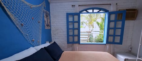 A room with a bed, a hammock, a window with blue shutters, and a view of greenery outside.