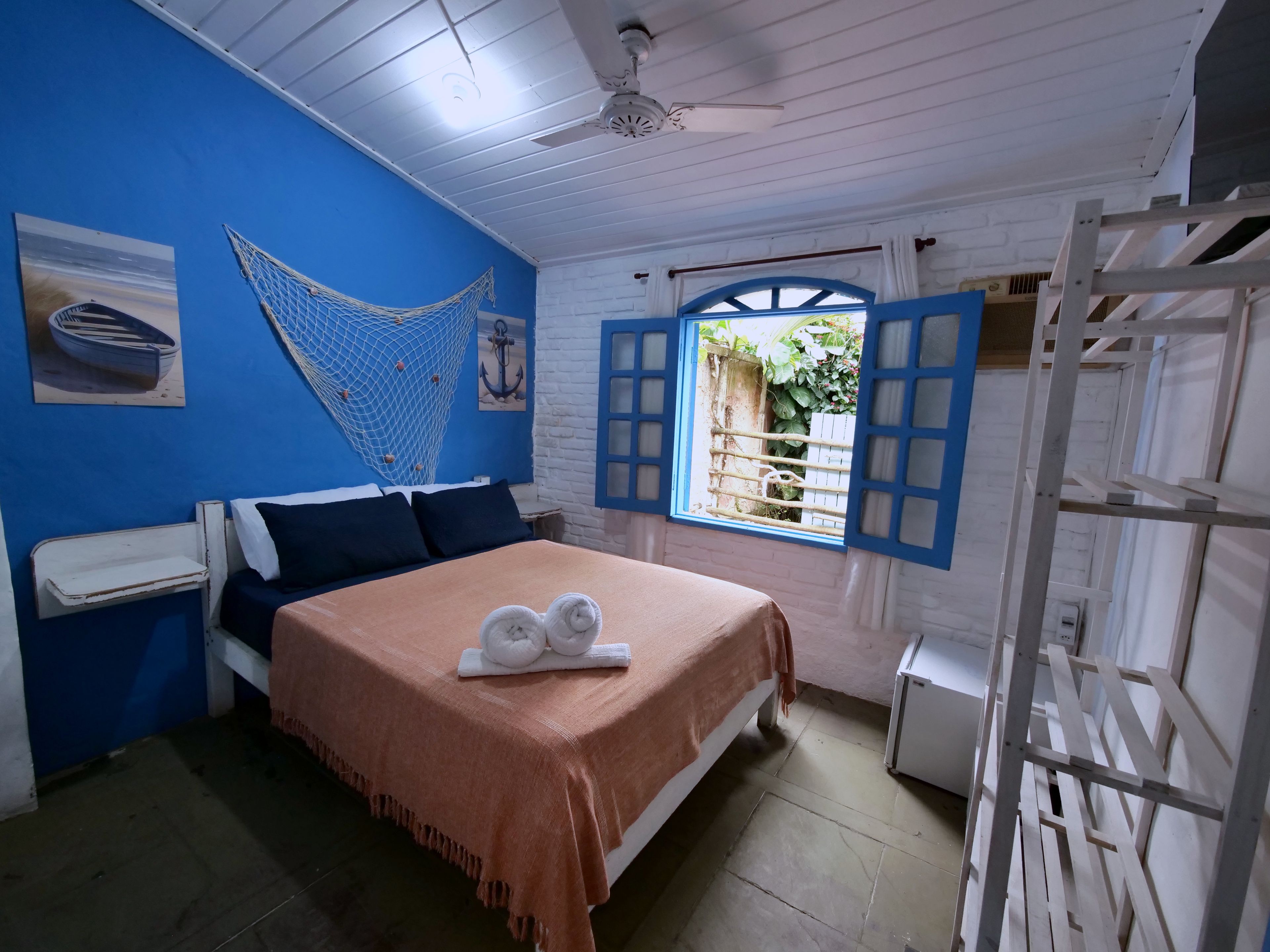 Ein Zimmer mit einem Bett, einem Deckenventilator, einem Fenster mit blauen Lamellen und einer Hängematte.