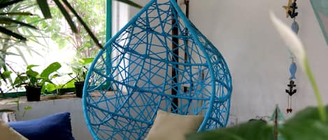 A hanging chair with a geometric pattern, a blue sofa, and potted plants in a room with a white wall and a window.