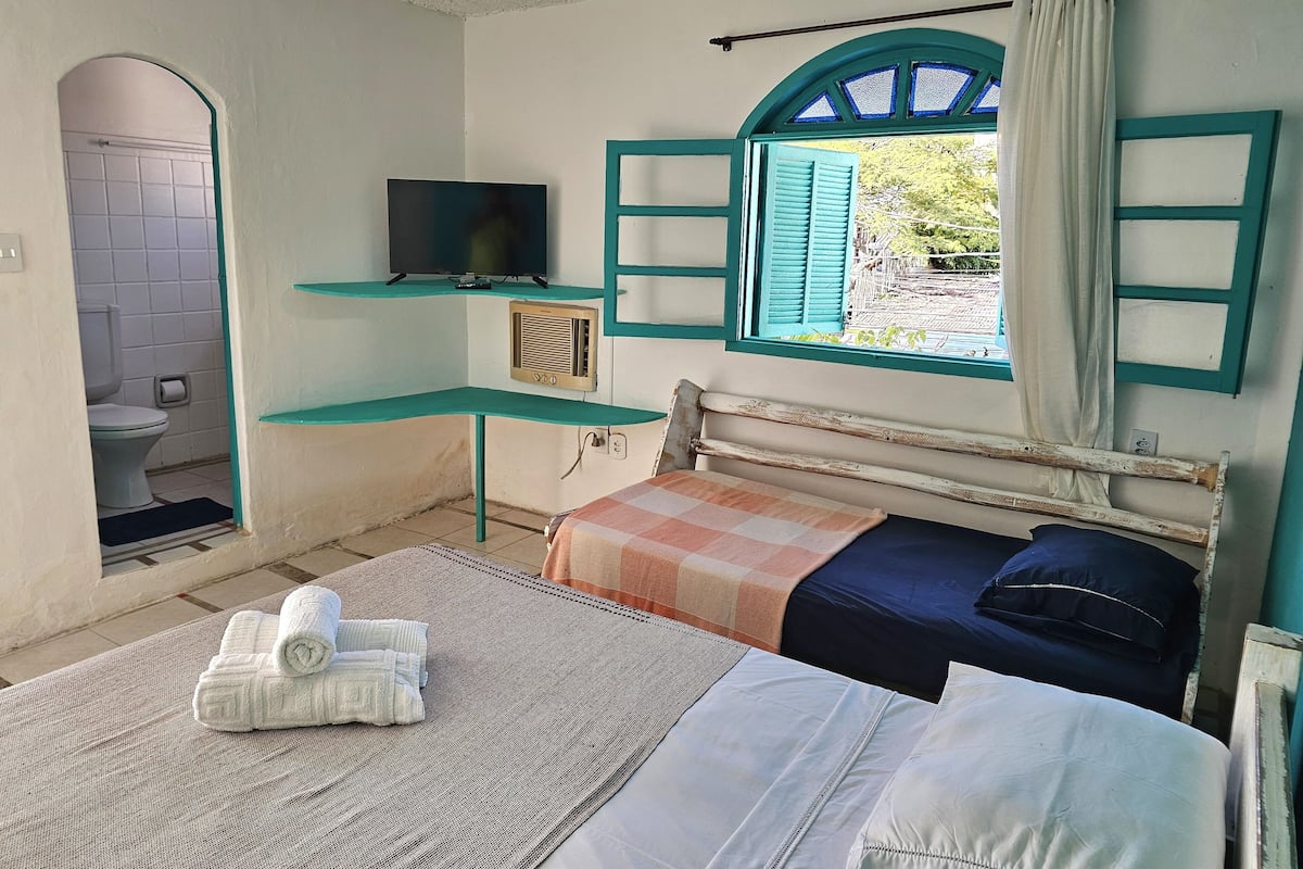 Ein Schlafzimmer mit einem Bett, einem Fernseher, einem Fenster mit Fensterläden und einem durch einen Bogen sichtbaren Badezimmer.