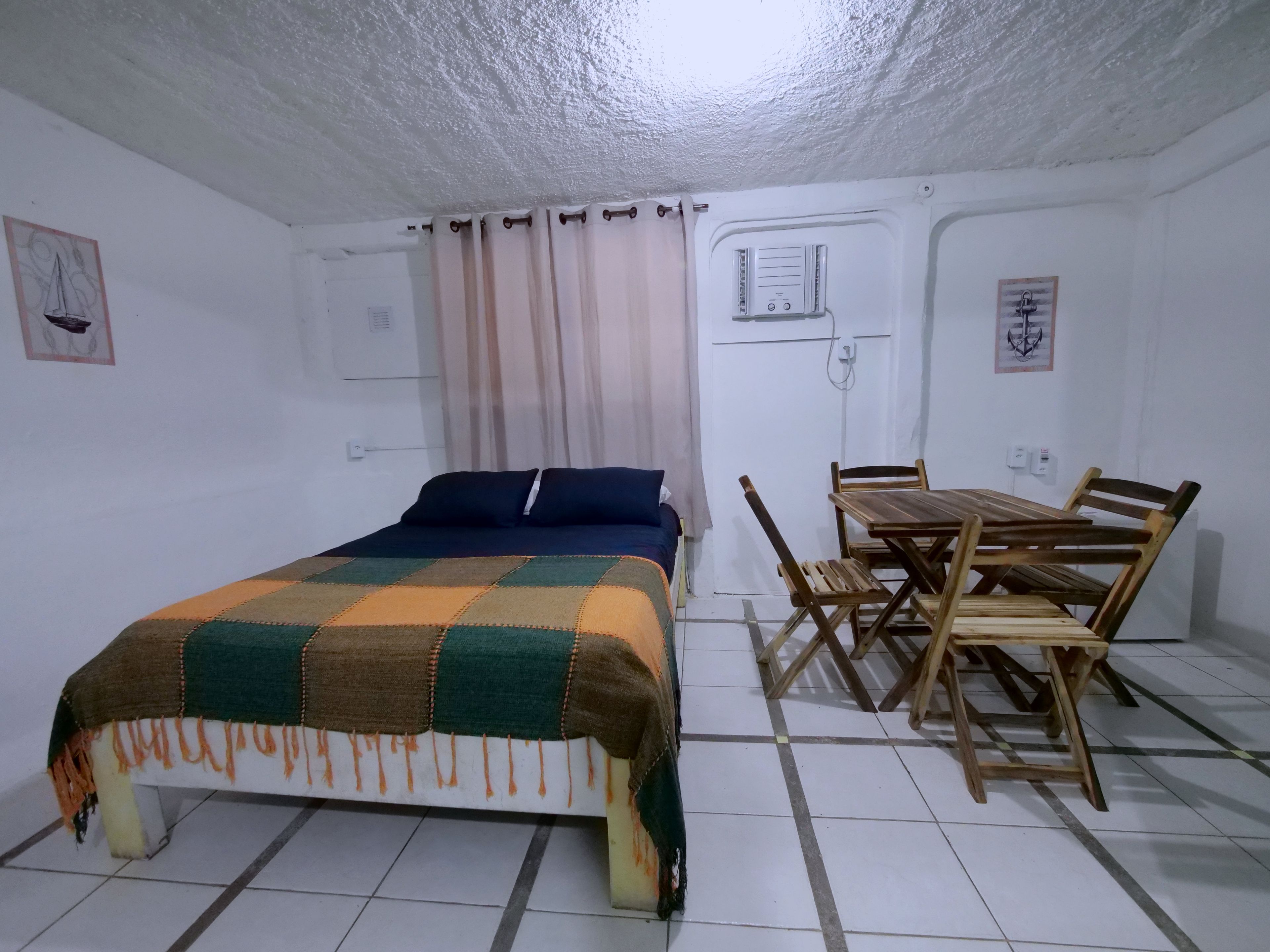 Une pièce comprenant un lit, une table à manger en bois avec des chaises, une fenêtre avec des rideaux et un climatiseur fixé au mur.