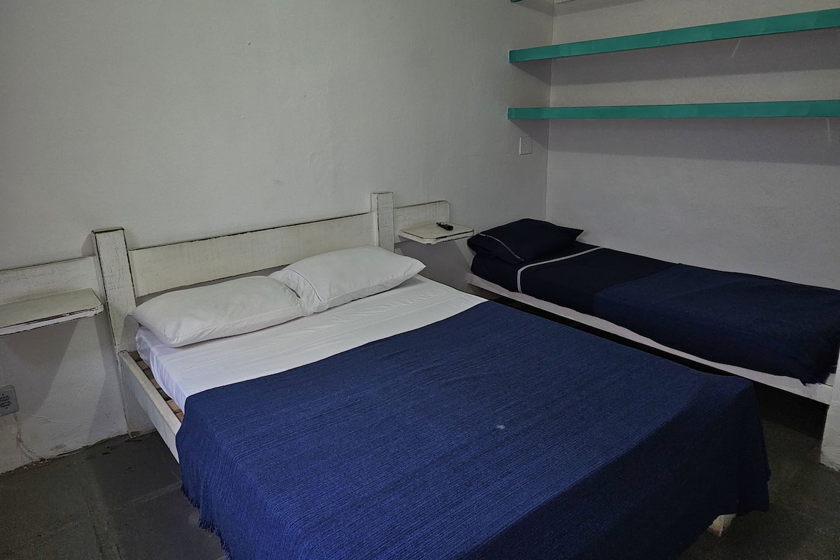 Ein Zimmer mit einem Einzelbett, einem weißen Kopfteil, einer blauen Bettdecke und einem weißen Kissen. Es gibt einen kleinen Nachttisch mit einer Fernbedienung.