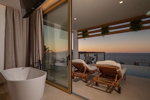 Een balkon met twee houten lounge fauteuils, een glazen deur en uitzicht op zee bij zonsondergang.