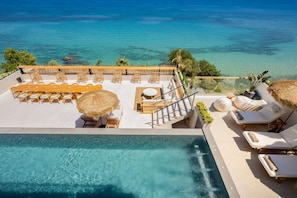 Zona de piscina en la azotea con sillones de descanso, una mesa de comedor y una escalera que lleva a un balcón con vistas al mar.