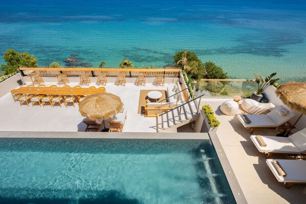 A rooftop pool area with lounge chairs, a dining table, and a staircase leading to a balcony overlooking the sea.