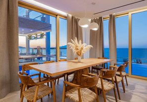 A dining area with a large wooden table and chairs, a vase with dried flowers, and a view of the sea through large windows.