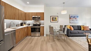 A modern kitchen with stainless steel appliances, wooden cabinets, and a dining area with a gray sofa and a glass table. - Comfortable 2BR ! Free parking ! 30 min to NYC ! (Newark)