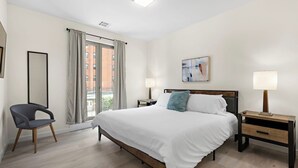A hotel room with a bed, two bedside tables, a chair, a large window with curtains, a painting on the wall, and a ceiling light. - Comfortable 2BR ! Free parking ! 30 min to NYC ! (Newark)