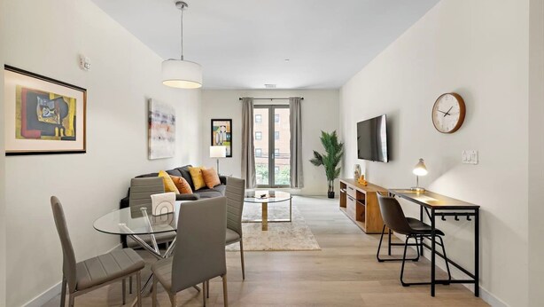 A modern living room with a sofa, coffee table, and dining area with chairs and a table. A TV and clock are mounted on the wall. - Comfortable 2BR ! Free parking ! 30 min to NYC ! (Newark)