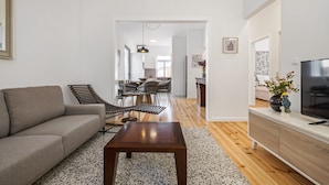 A modern living room with a sofa, a coffee table, and a TV on a stand. There is a dining area in the background with a table and chairs. - The Graceful Manor by Homie (Funchal)