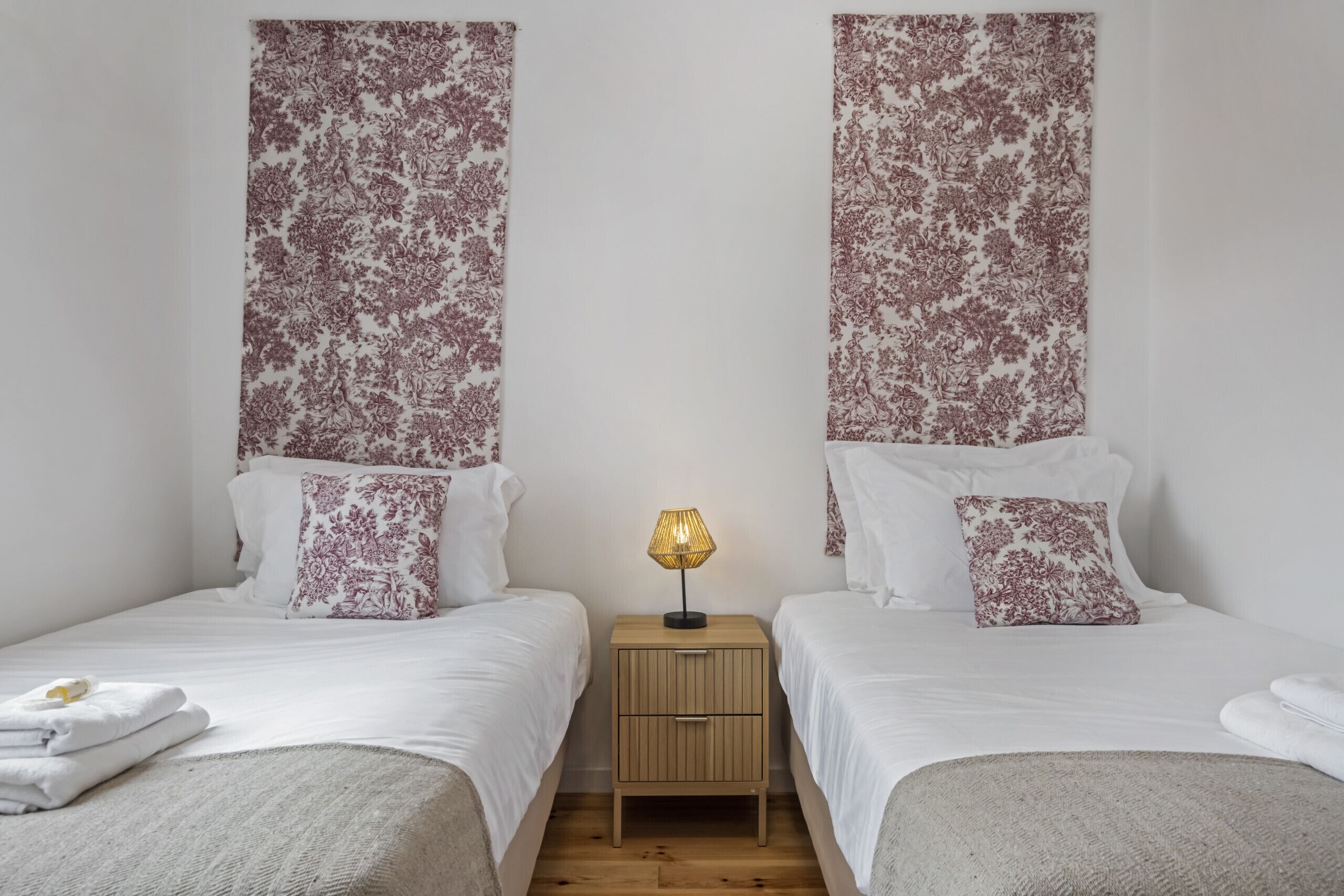 Ein Hotelzimmer mit zwei Betten, jedes mit weißer Bettwäsche und einem Kopfstück mit Blumenmuster. Zwischen den Betten steht ein kleiner Nachttisch mit einer Lampe.