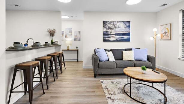 Un salon moderne avec un canapé gris, des tabourets en bois, une table basse et un tableau accroché au mur.