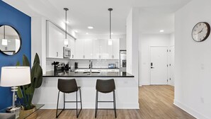 Una cocina moderna con armarios blancos, una isla central con dos taburetes de bar, un espejo redondo y un reloj en la pared.