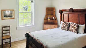 Ein Schlafzimmer mit einem großen Bett, einem hölzernen Nachttisch, einem Sessel, einem Fenster mit Blick auf Bäume und einem Bild an der Wand.