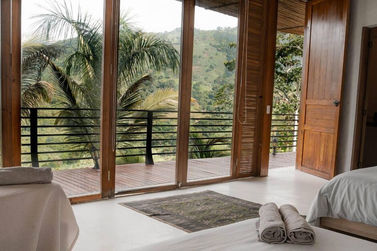 Ein Schlafzimmer mit großem Fenster, einer Holzterrasse und Blick auf eine üppige Landschaft.