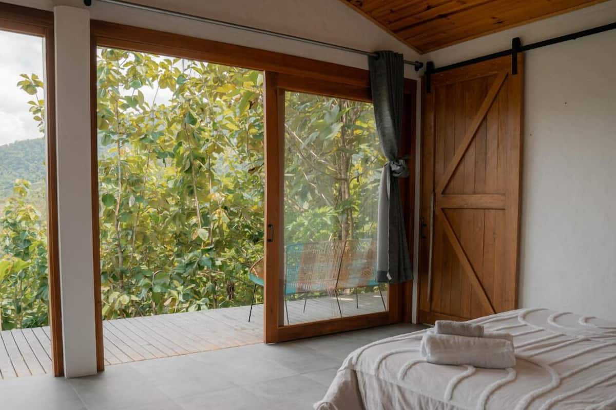 Ein Schlafzimmer mit einem großen Bett, einer Schiebetür, die auf einen Balkon führt, und Blick auf Grünflächen.