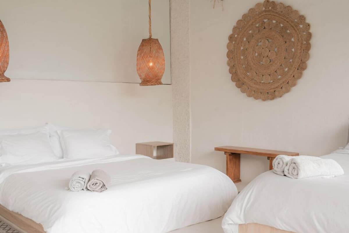 Ein schlicht eingerichtetes Schlafzimmer mit zwei Betten, einem Holznachttisch, einer gewebten Wanddekoration und einer Pendelleuchte.