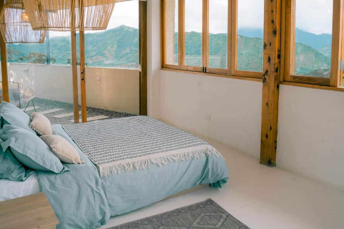 Ein Schlafzimmer mit Bett, einer Pendelleuchte, großen Fenstern und Blick auf Berge.