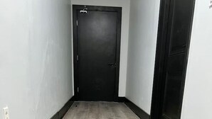 Un pasillo estrecho con suelo de madera, una puerta negra al final y una pared blanca con un interruptor de luz a la izquierda.