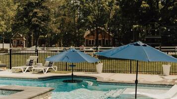 Piscina con sillas de descanso y sombrillas, rodeada de una valla y árboles.
