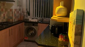 A compact kitchen with a gas stove, a washing machine, and a sink. There is a window with vertical blinds, a yellow vase on a shelf, and a painting on the wall. - Komegloba Exquisite room (Manchester)