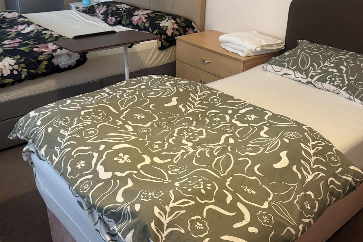 A hotel room with two beds, each with floral-patterned bedding, a bedside table with a lamp, and a small desk with a lamp and papers.