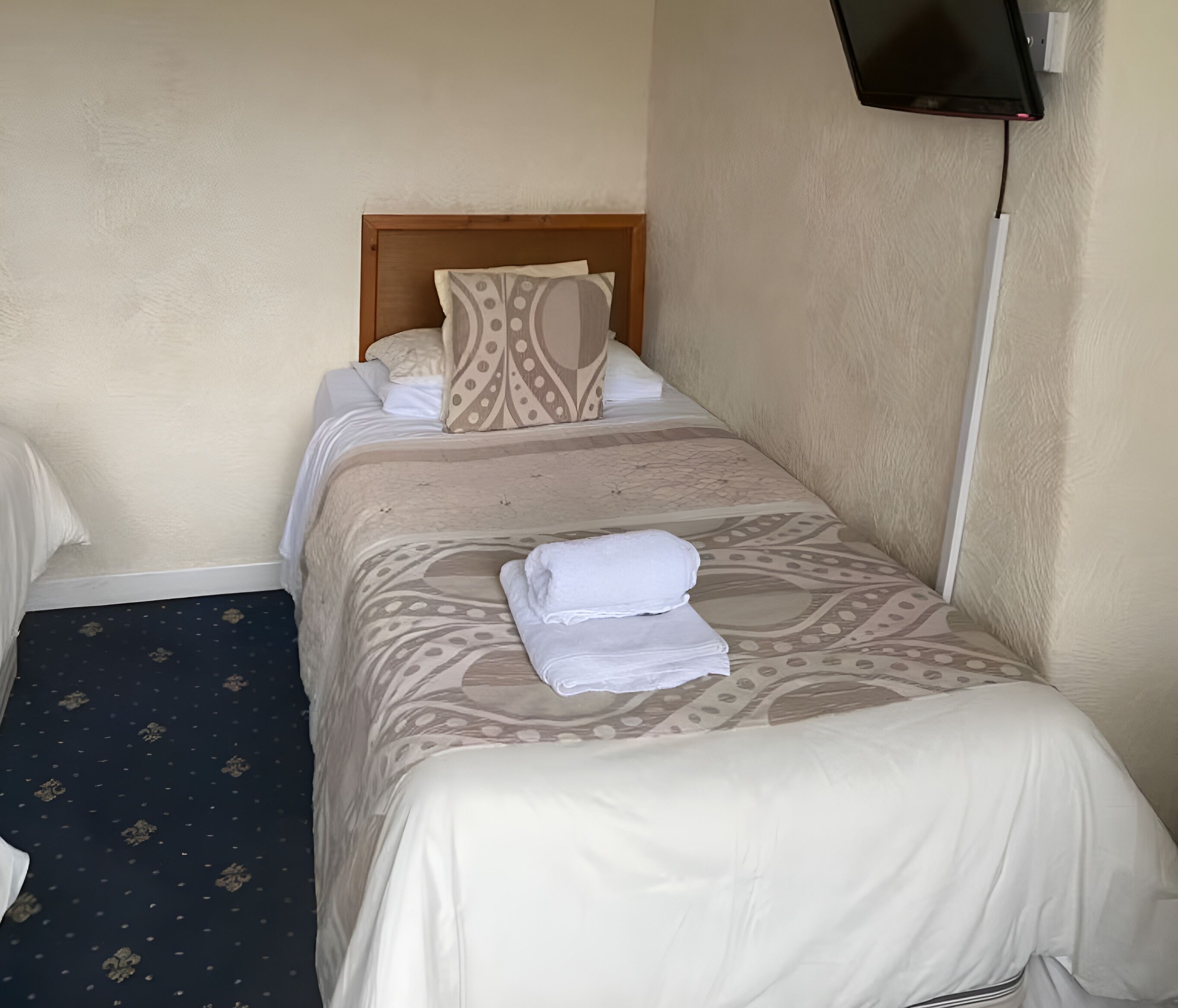 A neatly made bed with a patterned bedspread and pillows, a folded white towel on the bed, and a television mounted on the wall.