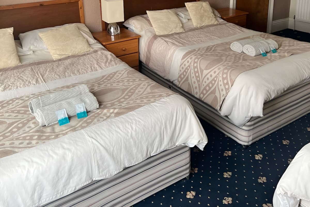 A hotel room with two beds, each with a wooden headboard, a bedside table with a lamp, and a rolled towel on the bed.