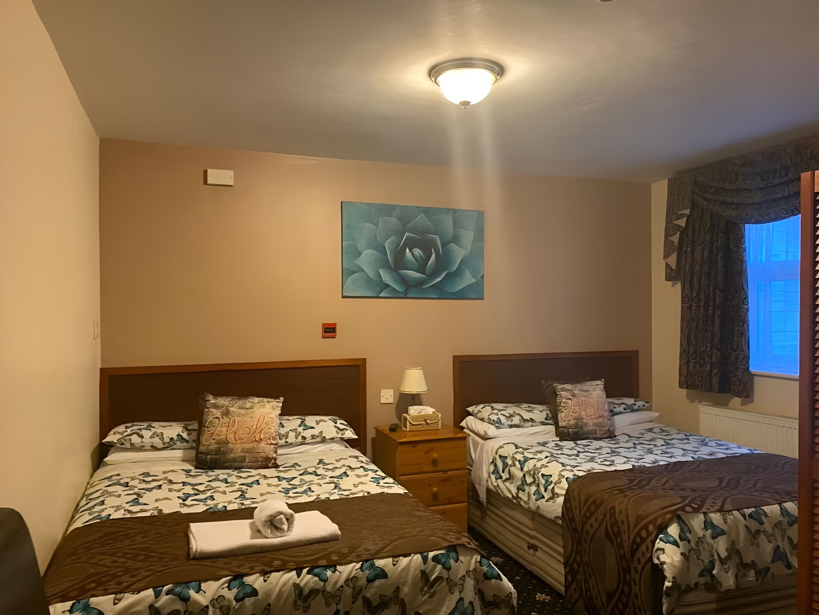 A hotel room with two beds, a nightstand, a lamp, a flower painting, and a window with curtains.