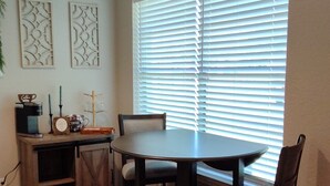 Un comedor con una mesa redonda y cuatro sillas, una estación de café y una ventana con persianas.