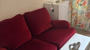 A red sofa, a white side table, and a small white cabinet with a floral curtain in the background. - Room with private bathroom, MOULIN ROUGE, 20 mn from PARIS SHARED APARTMENT (Épinay-sur-Seine)