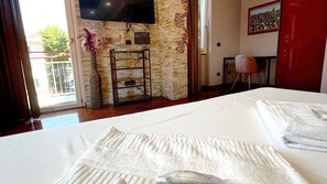 Una camera d'albergo con un letto, una televisione fissata a una parete in pietra, un tavolino con una sedia e una finestra vista verde.