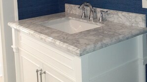 Un baño con tocador blanco, encimera de mármol, espejo redondo con marco de fibra natural y pared de textura azul.