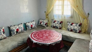 Une pièce avec une table ronde, des canapés de couleur beige, des coussins à motifs et une fenêtre ornée de rideaux légers.