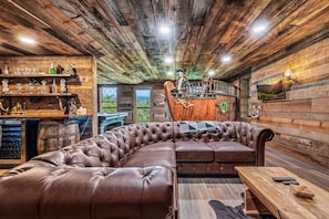 A rustic cabin interior with a leather sectional sofa, wooden walls, a bar area with bottles and glasses, and a scenic view through a door. - INACTIVE - Ritz of The Smokies (Gatlinburg)