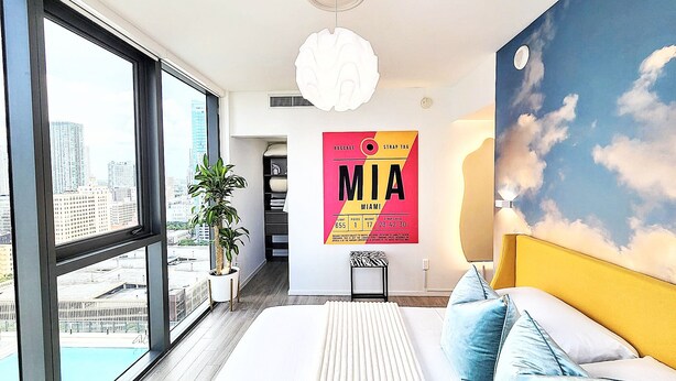 A modern bedroom with a large window, a city view, a bed with blue pillows, and a 'MIA' poster on the wall. - NEW! Electric Luxury Downtown Bayfront Brickell (Miami)