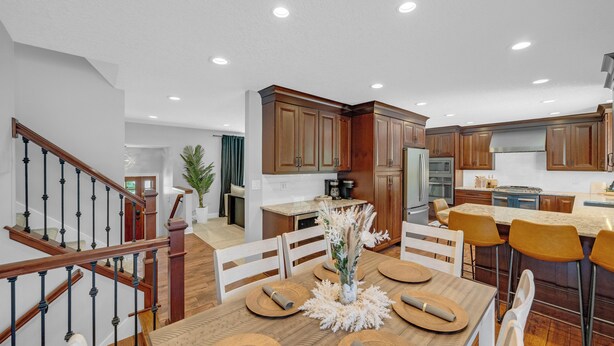 A modern kitchen with wooden cabinets, a stainless steel refrigerator, and a dining area with a wooden table and chairs. - Urbandale Oasis (Urbandale)