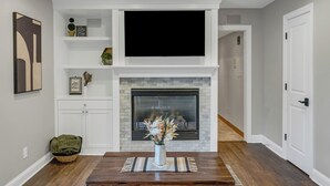 A living room with a fireplace, a flat-screen TV mounted above it, and a dining table with a vase of flowers. - Urbandale Oasis (Urbandale)